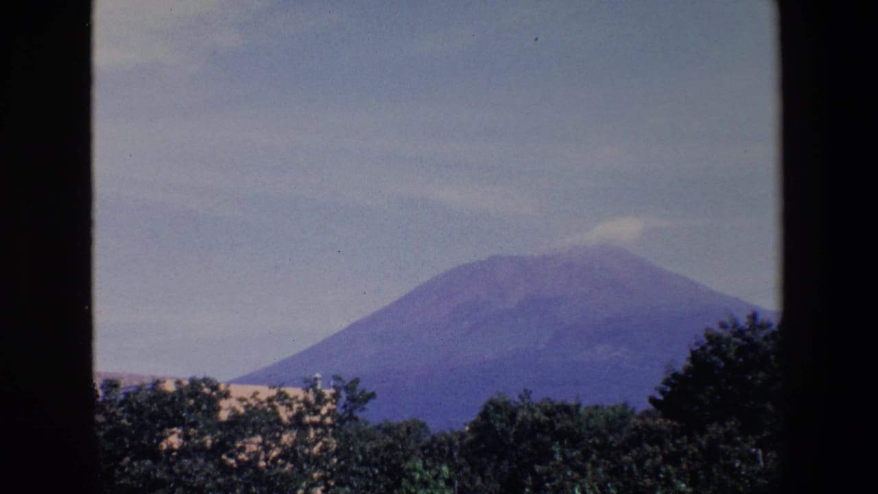 Distant view of a broad mountain identified in the clip metadata as Mount Etna, with hazy sky and treetops in a 1967 Sicily home-movie shot.