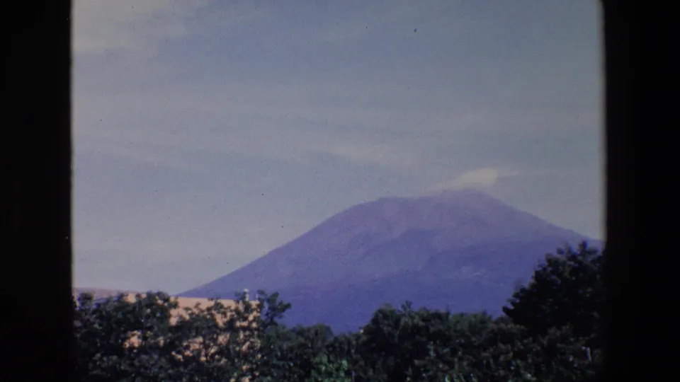 Distant view of a broad mountain identified in the clip metadata as Mount Etna, with hazy sky and treetops in a 1967 Sicily home-movie shot.