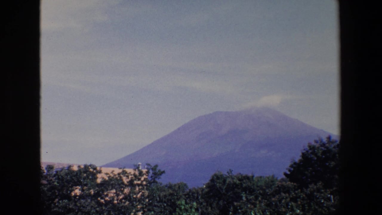 1967 Palermo home-movie frame showing a distant conical mountain beneath a pale blue sky, with dark window framing and trees in the foreground.