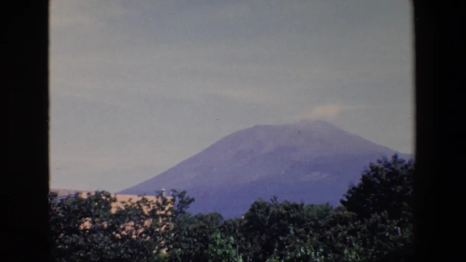 1967 Palermo home-movie frame showing a softly focused mountain view through dark framing, with trees and light rooflines in the lower foreground.