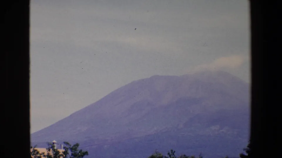 1967 Palermo home-movie frame showing a closer view of the mountain slope under a pale sky, with a faint summit cloud or plume and visible film dust or scratch marks.