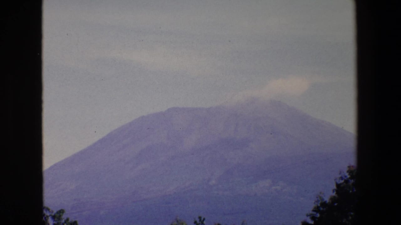 1967 Palermo home-movie frame with a distant mountain under a hazy sky and a light plume near the summit, bordered by dark window framing.