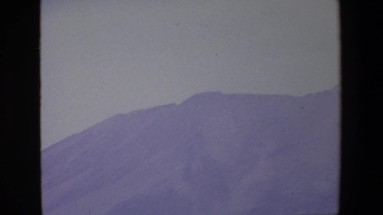 1967 Palermo home-movie frame showing a hazy blue-purple mountain slope viewed through a dark window frame under a pale sky.