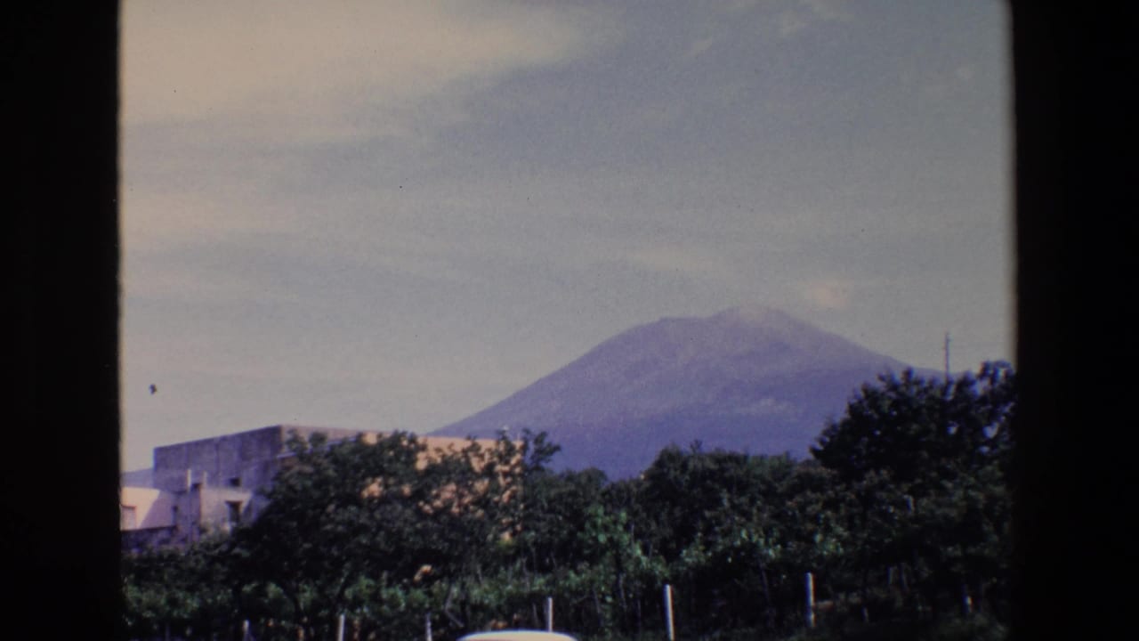 Distant view of Mount Vesuvius above trees and low buildings in 1967 Italian 8mm travel footage.