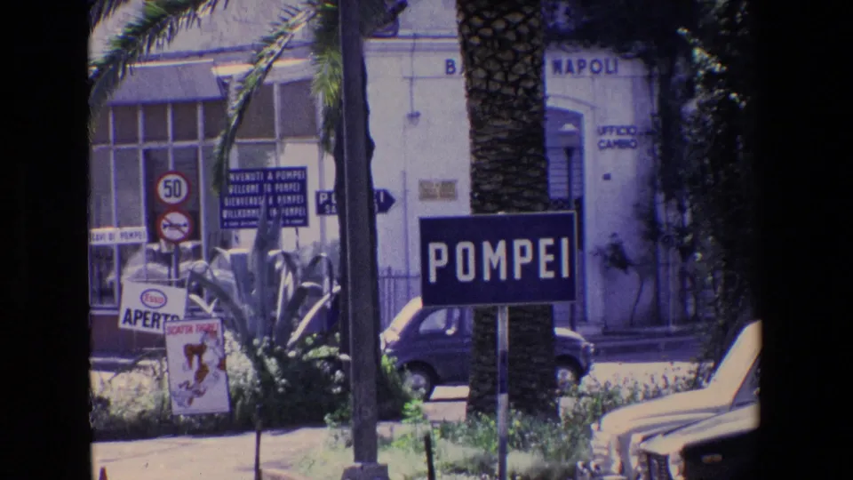 Street-level Pompeii view from 1967 home-movie footage, showing a blue POMPEI sign, palm tree, compact cars, road signs, and building signage with NAPOLI lettering.