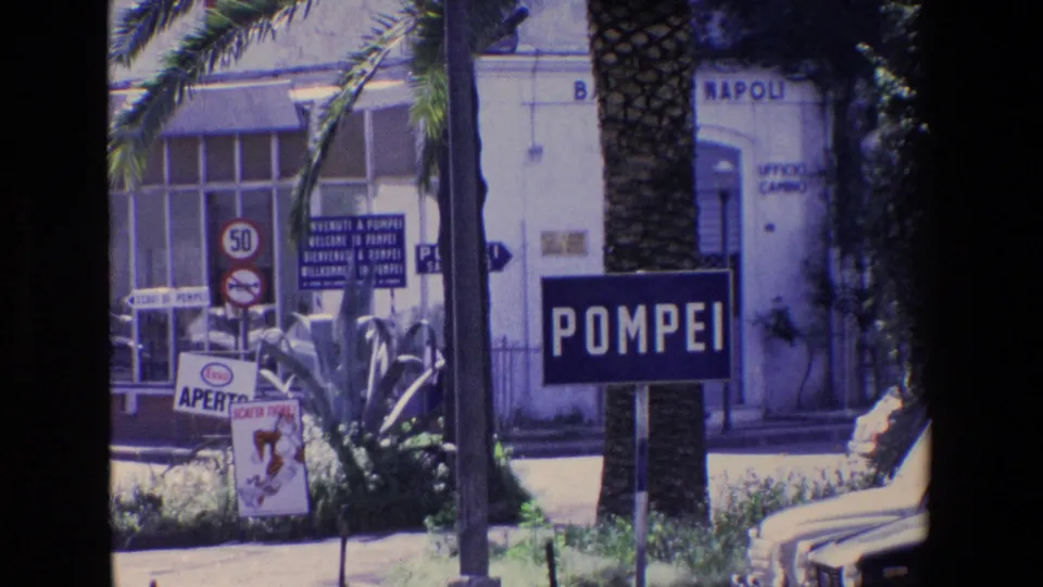 Pompeii street scene with the blue POMPEI sign prominent in the foreground, a palm trunk, road signs, compact cars, and NAPOLI lettering on a nearby building.