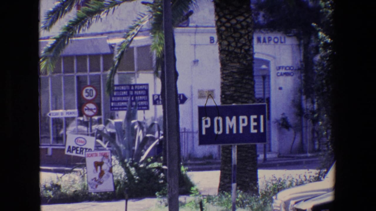 1967 Italian travel footage showing a street approach to Pompeii with a prominent POMPEI sign, palm trunks, road signage, and a building marked with Napoli text.