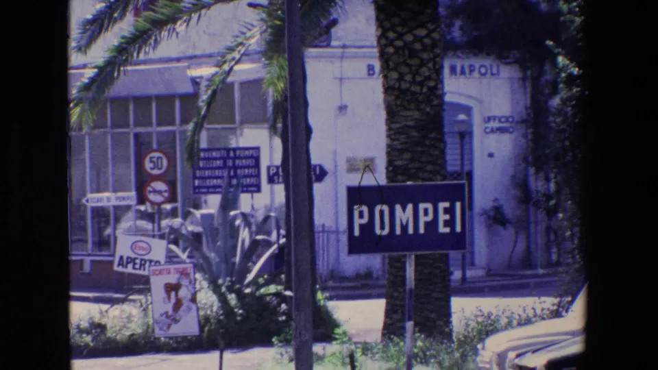 1967 Italian travel footage showing a street approach to Pompeii with a prominent POMPEI sign, palm trunks, road signage, and a building marked with Napoli text.