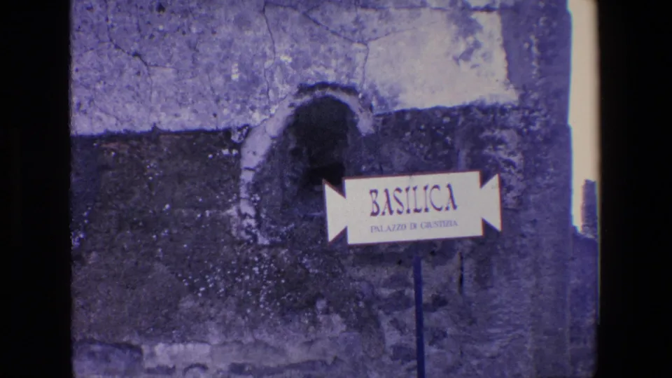 Visitor-shot 8mm frame of the Basilica sign at Pompeii, held close on the signpost and surrounding aged masonry.