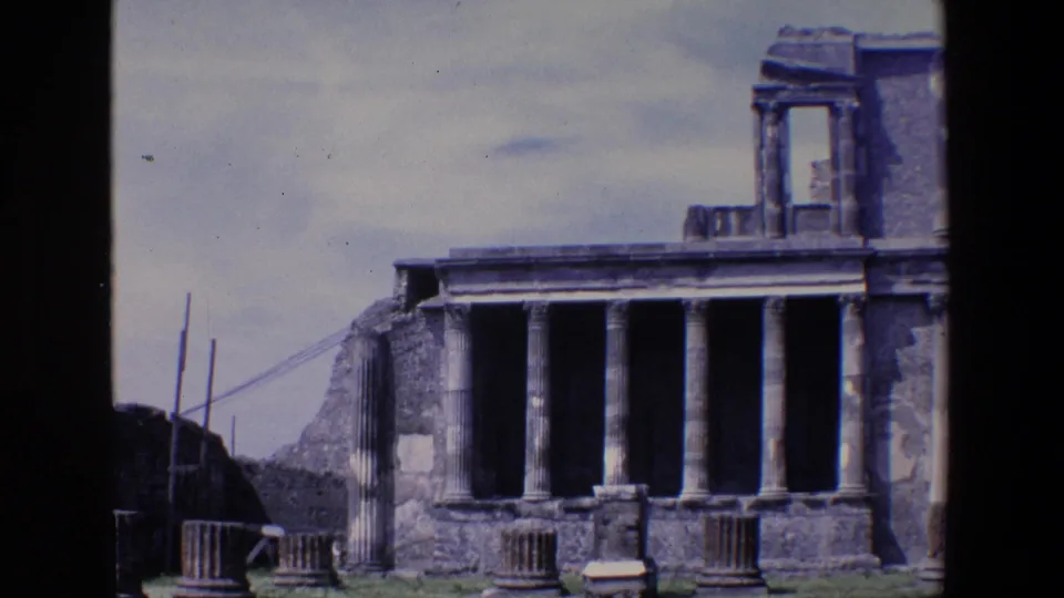1967 home-movie view of Pompeii ruins showing weathered columns, stone fragments, and a classical structure framed from a visitor vantage point.