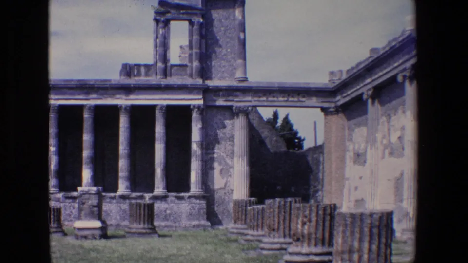 1967 Palermo frame showing the columned ruin from a slightly shifted angle, with an adjoining wall at right, grass in front, scattered stone pieces, and pale daylight sky.