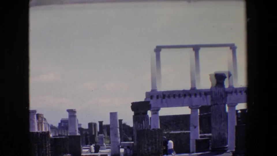 1967 Palermo home-movie frame with a slightly tilted view of standing columns, dark ruin walls, low stone remains, and a small visitor near the lower foreground.