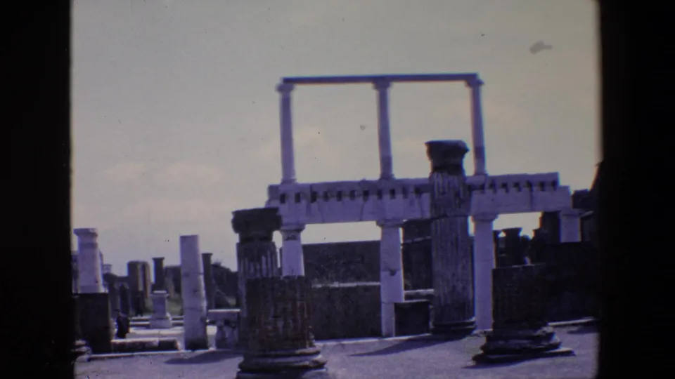 1967 Palermo frame showing classical stone ruins with upright columns, block walls, a paved path, and distant visitors under a pale sky.