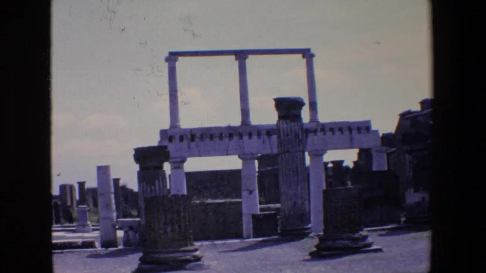 1967 Palermo, Italy home-movie frame showing weathered stone columns, low ruin walls, and a pale overhead architectural structure against a bright sky.