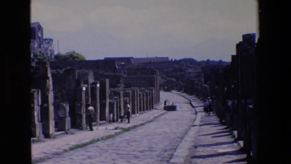 1967 Palermo, Italy home-movie frame looking along a cobblestone street through a historic ruin area, with old stone structures, shadows, and a few distant people.