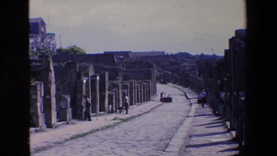 1967 Palermo, Italy home-movie frame showing a cobblestone street bordered by weathered stone ruins, with several distant visitors walking through the site.