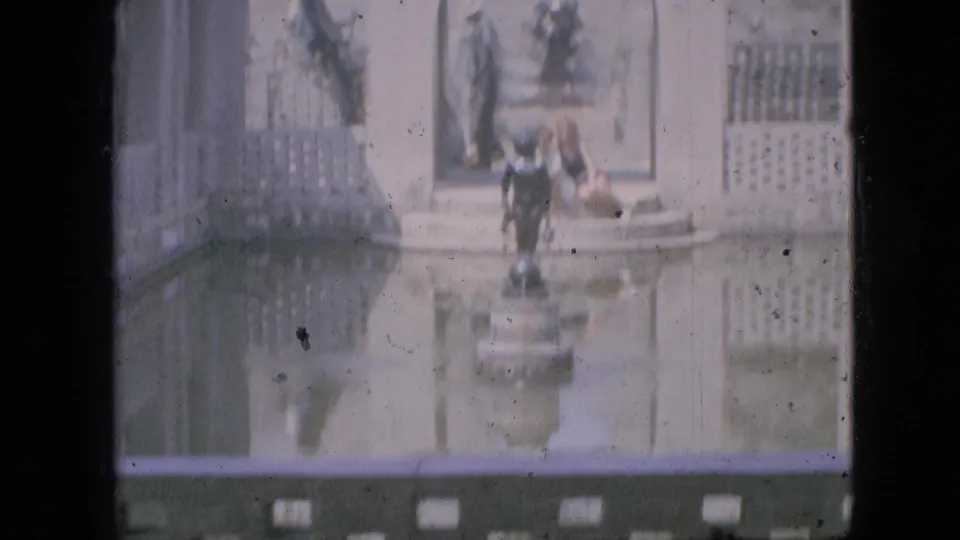 Faded 8mm frame from Branson, Missouri in 1968 showing a central fountain statue, reflective water, pale walls, lattice panels, and people gathered near the fountain edge.