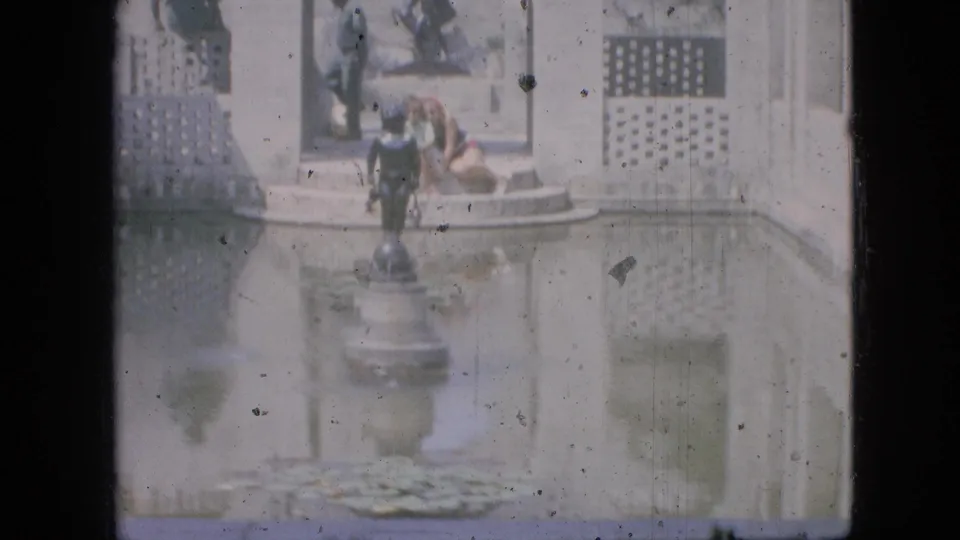 1968 Branson, Missouri home-movie still with a person seated on the fountain edge, a standing figure nearby, a central statue, lattice walls, and sunlit water reflections.