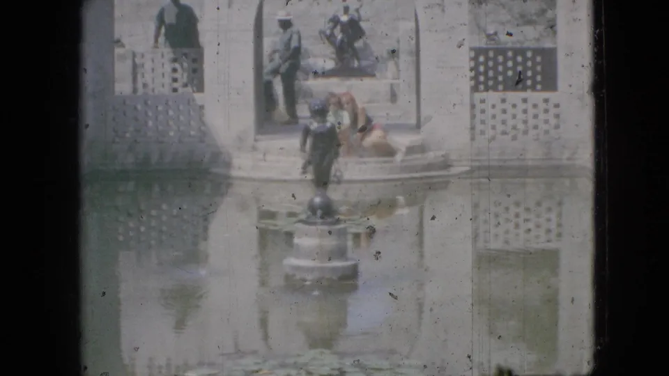 Soft 8mm frame from the stated 1968 Branson location showing a person seated near the fountain, another figure standing on the steps, a small statue, latticework, and reflective water.