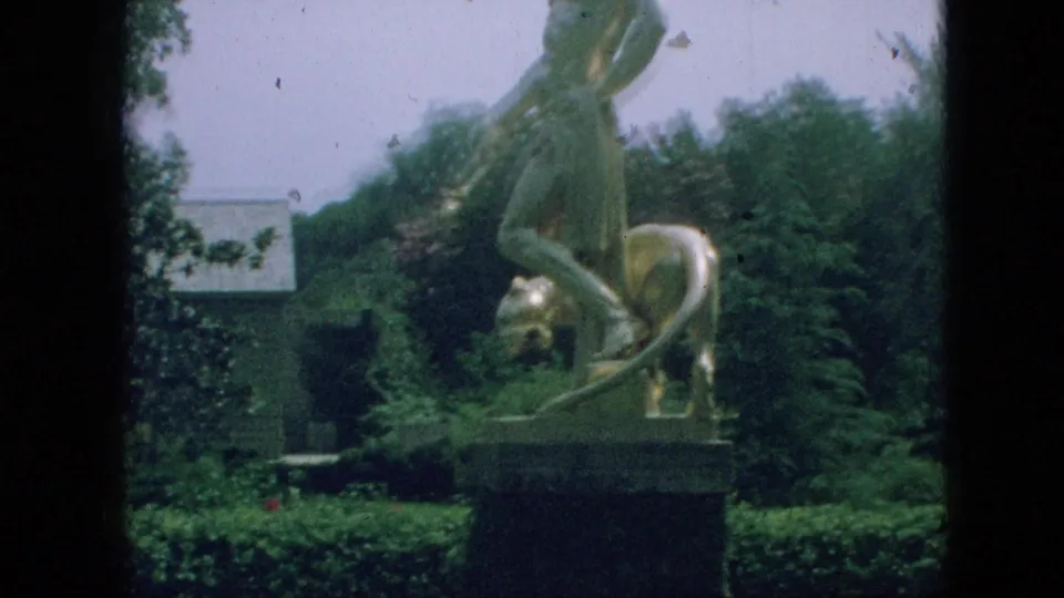 Wider 1968 Branson home-movie view of the Adam and Eve with dog sculpture on a pedestal in a garden, with hedges, trees, and a low building in the background.