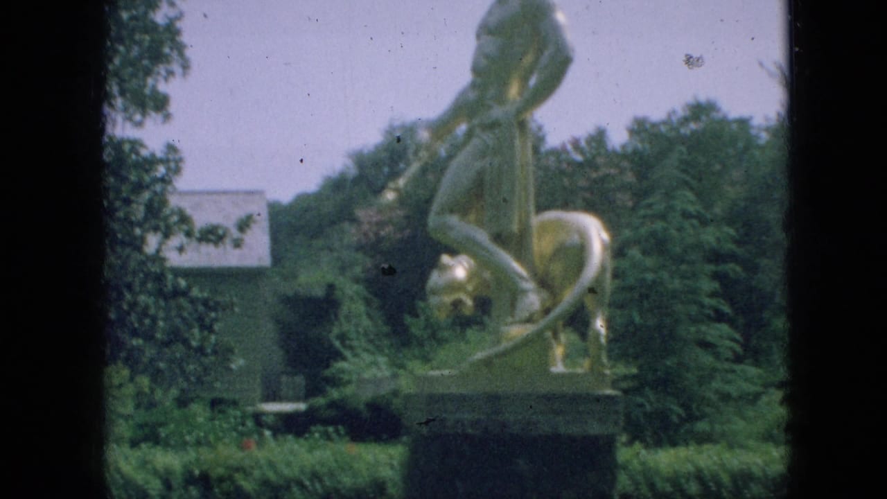 1968 Branson, Missouri home-movie frame showing a gold-toned figure-and-lion statue on a pedestal amid hedges, trees, lawn, and a pale background building.