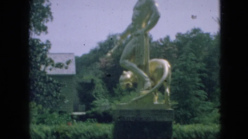 1968 Branson, Missouri home-movie frame showing a gold-toned figure-and-lion statue on a pedestal amid hedges, trees, lawn, and a pale background building.