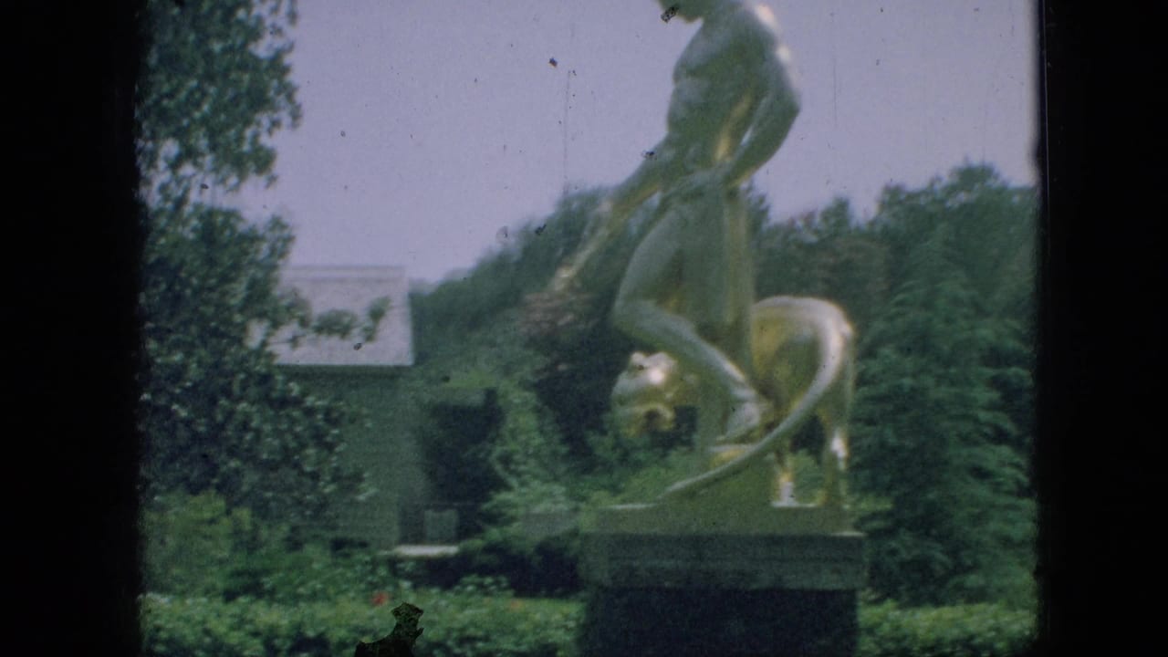 Gold-toned statue and pedestal in a landscaped garden with trees and hedges.