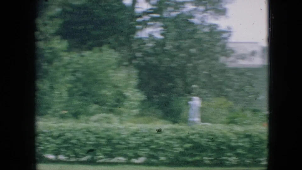 Motion-blurred 1968 Branson, Missouri home-movie frame showing garden greenery, clipped hedges, lawn, trees, and a pale building as the camera moves away from the statue.