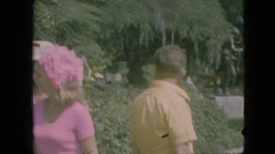 Two adult park visitors, one in a pink top near pink flowers and one in a yellow shirt, walk beside dense greenery in the Branson park footage.