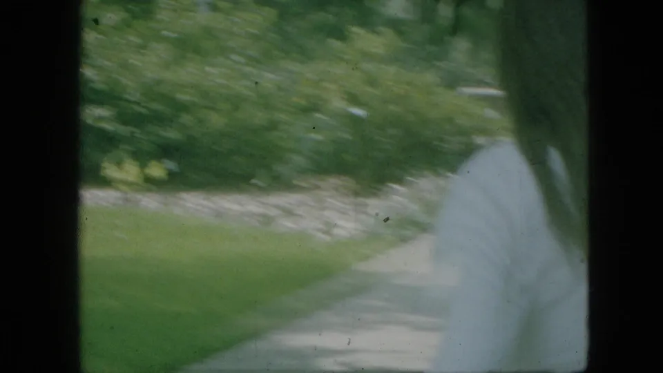 A woman in a light dress appears in motion on a paved path in a landscaped Branson, Missouri park scene dated 1968 by metadata, with grass, trees, and planting beds nearby.