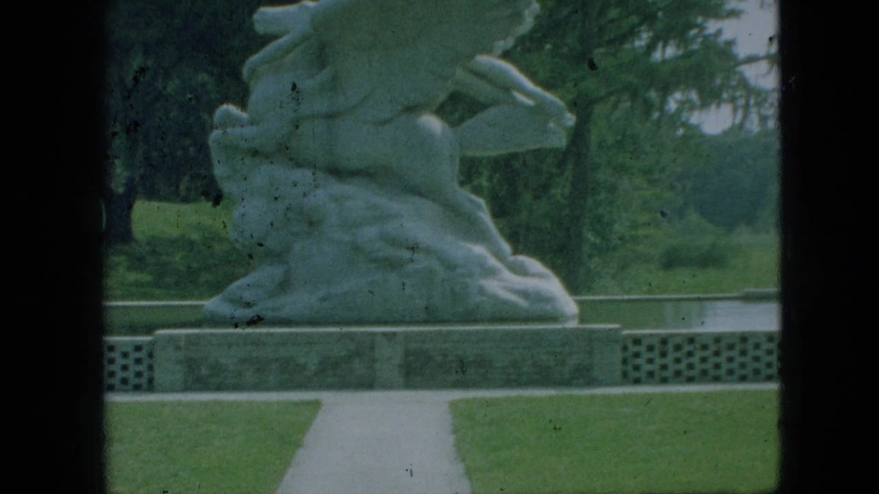 1968 Branson, Missouri 8mm frame showing a pale winged stone sculpture on a plinth, bordered by a straight walkway, green lawn, and trees.