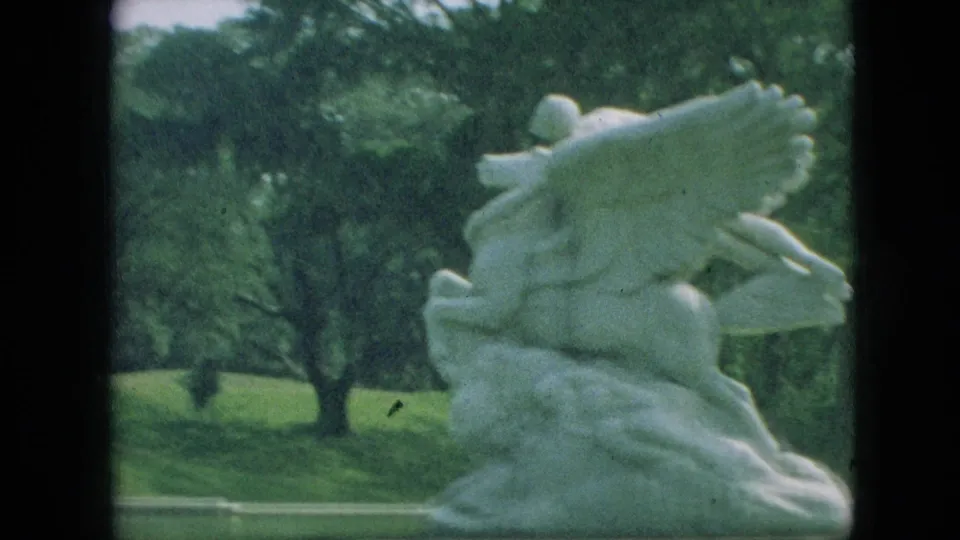1968 Branson, Missouri 8mm frame showing the pale winged sculpture from a slightly shifted angle, with trees and lawn behind it.