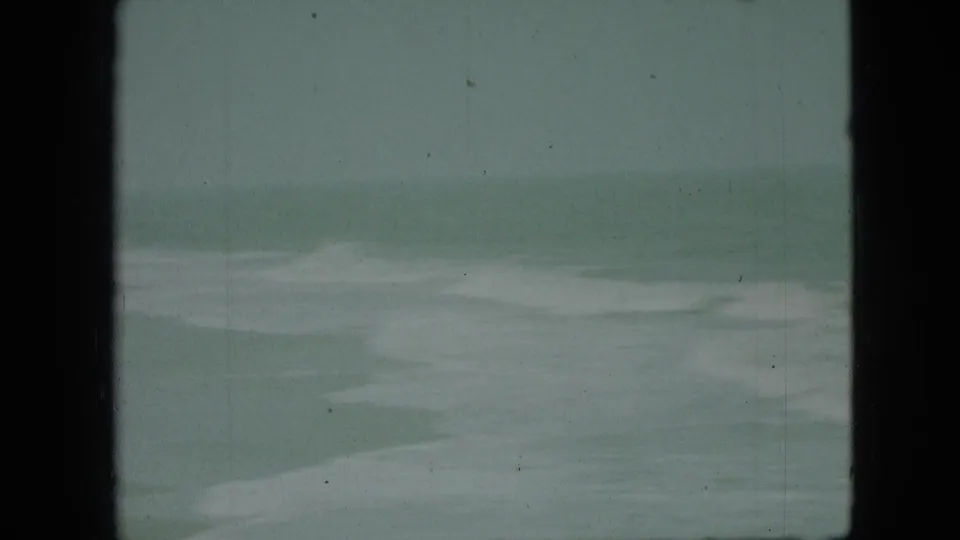 Green-blue 8mm still from the Branson vacation footage showing pale wave shapes, soft focus, and a dark vertical frame edge.