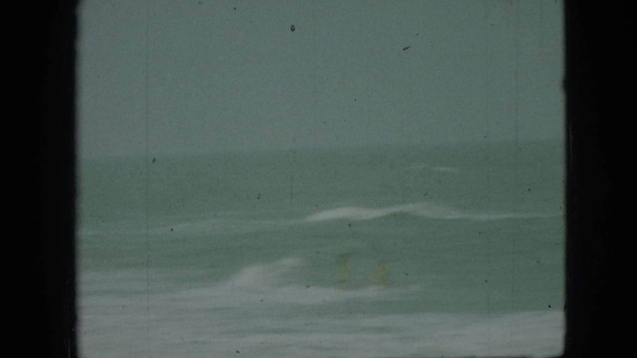 1968 8mm home-movie frame with greenish water, pale surf, and dark vertical masking or obstruction at the sides.