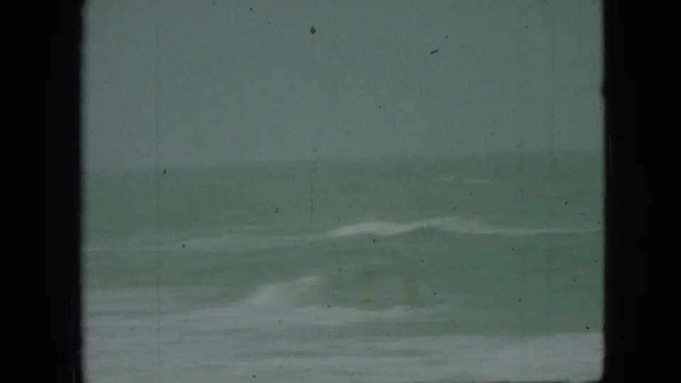 1968 8mm home-movie frame with greenish water, pale surf, and dark vertical masking or obstruction at the sides.