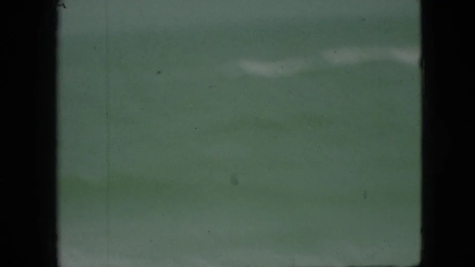Sparse shoreline view from the 1968 home-movie clip, showing muted water and a thin pale wave line with heavy dark framing at left.