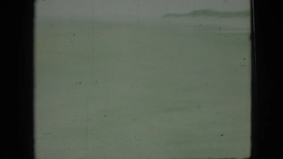 Wide, soft 8mm view of pale exposed sand or shallow water with low surf in the distance and dark in-camera frame borders.