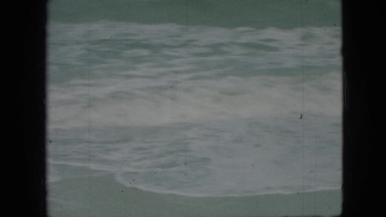 1960s 8mm home-movie frame of rough, foamy water seen from a beach or shoreline viewpoint, with visible gate edges and film wear.