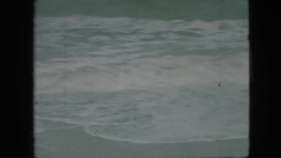 1960s 8mm home-movie frame of rough, foamy water seen from a beach or shoreline viewpoint, with visible gate edges and film wear.