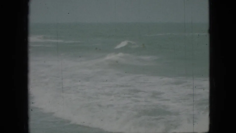Archival 8mm frame showing breaking surf and white foam under a dull sky, with scratches and dark frame edges visible in the scan.