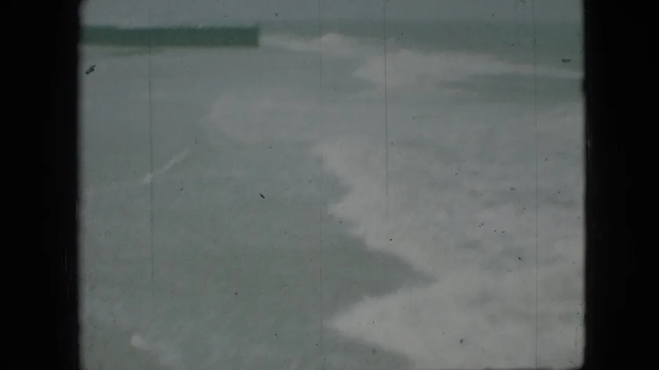 Soft, green-tinted 8mm frame of churning surf near a beach, with a low dark structure or breakwater visible across the upper left.