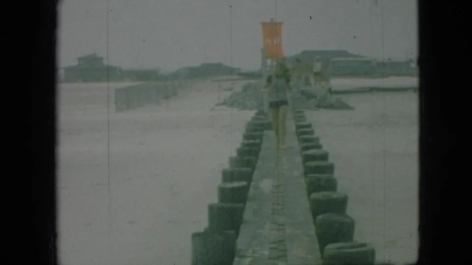 1960s 8mm frame showing a narrow beach jetty or walkway lined with posts, with a distant woman-like figure and an orange elevated structure near the shore; the supplied location is not visually confirmed.