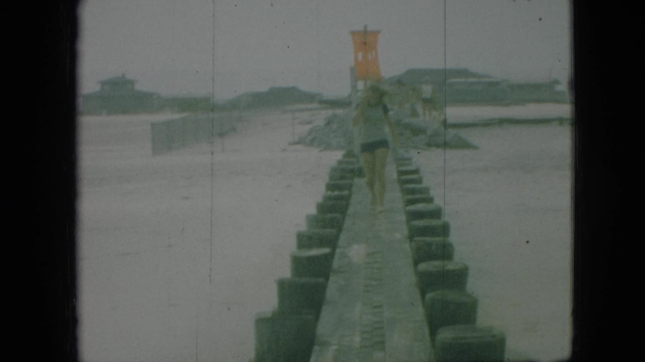 1968 Branson, Missouri 8mm frame showing a distant person walking along a narrow concrete jetty between rows of pilings near calm water.