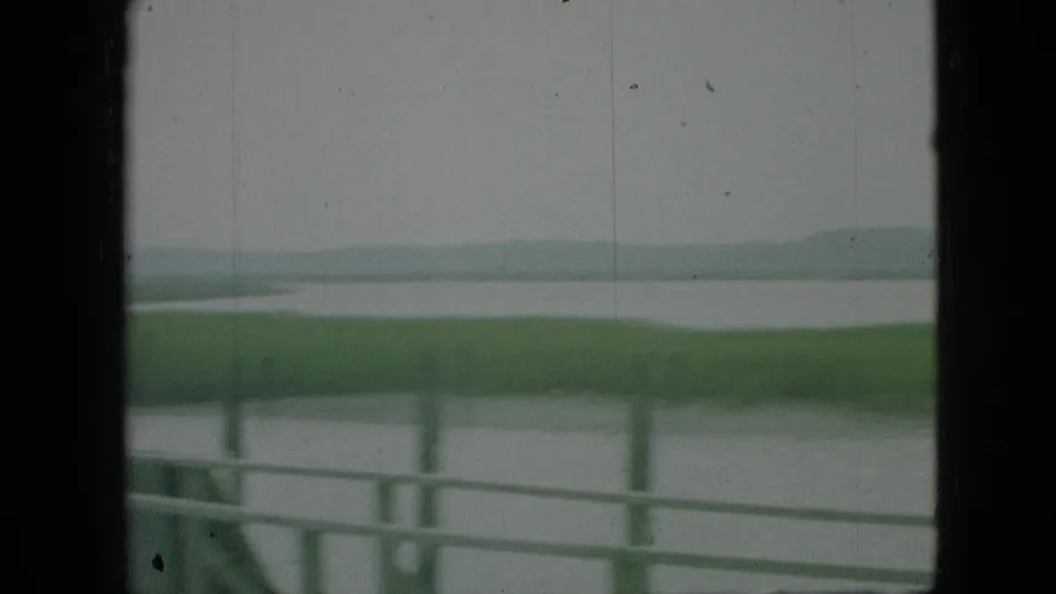 1968 Branson, Missouri 8mm frame looking past a foreground railing toward calm water, low marshland, and hazy distant hills.
