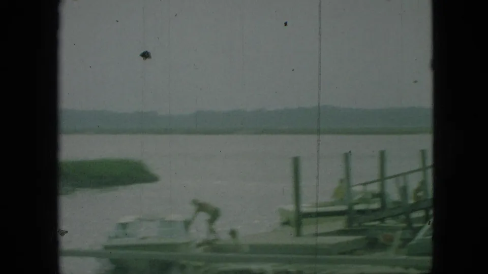1968 Branson, Missouri 8mm frame showing a small boat near wooden dock posts, with open water, green shore, and hazy hills in the background.