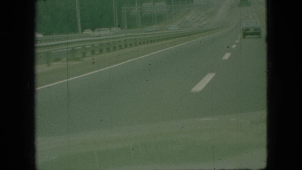 1968 Branson, Missouri 8mm home-movie frame showing a green-tinted highway view from a moving vehicle, with white lane markings, metal guardrails, and a distant car ahead.