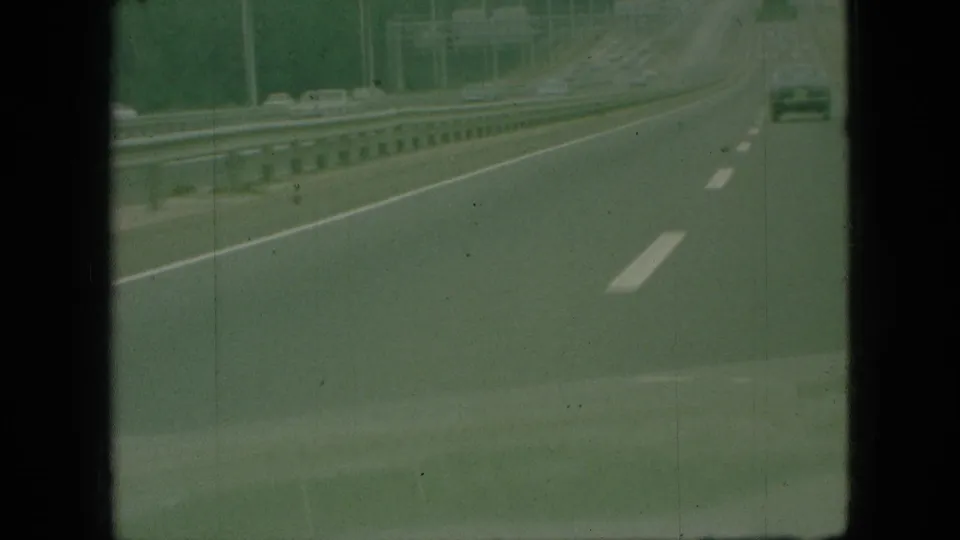 1968 Branson, Missouri 8mm home-movie frame showing a green-tinted highway view from a moving vehicle, with white lane markings, metal guardrails, and a distant car ahead.