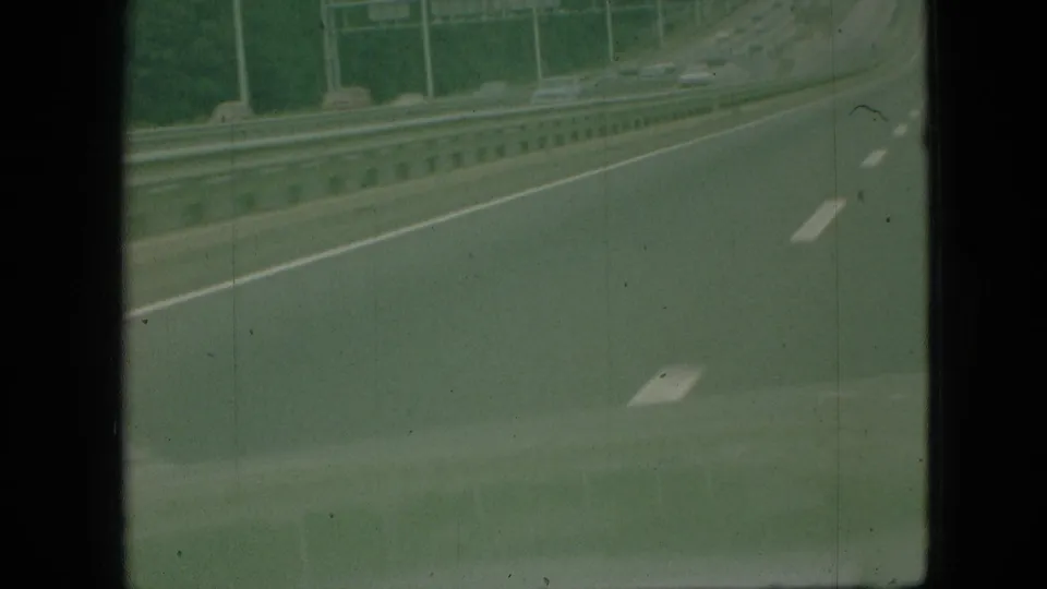1968 Branson, Missouri 8mm frame showing a moving-vehicle highway view with lane stripes, metal guardrail, roadside verticals, and visible film grain.