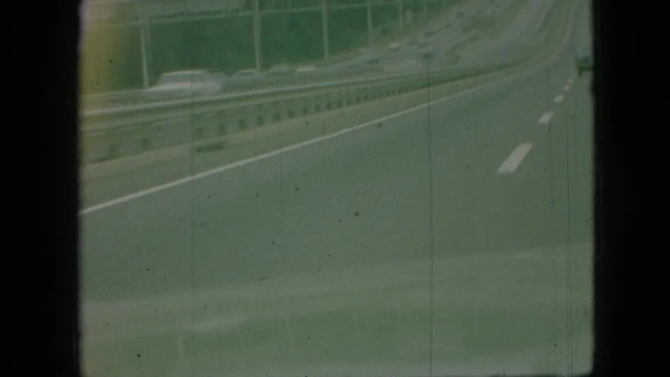 1968 Branson, Missouri home-movie frame showing a highway curve from a moving car, with guardrails, white lane markings, roadside posts, and faint distant traffic.