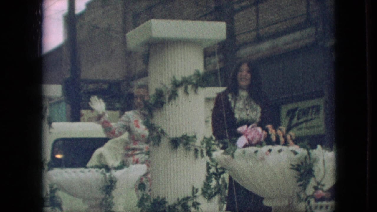 Two women ride on a garlanded parade float with a vehicle behind them.